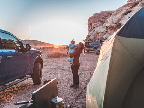 A woman holding a small child who is wearing a Morrison Outdoors sleeping Bag near a truck and a tent outdoors