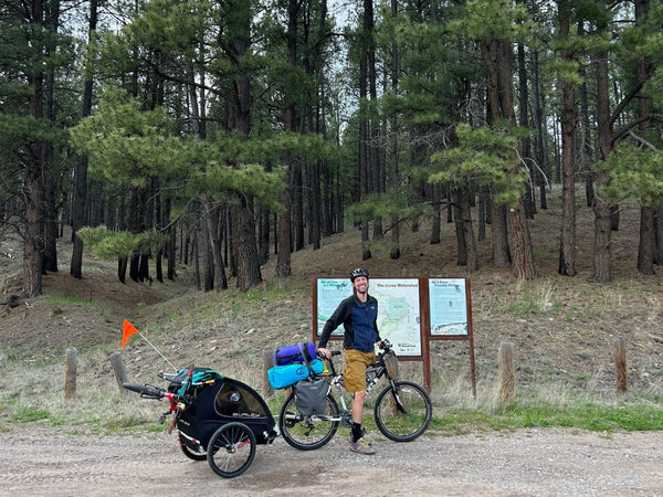 A man standing in front of a trail sign while on his bike that has a Burley trailer attached to it